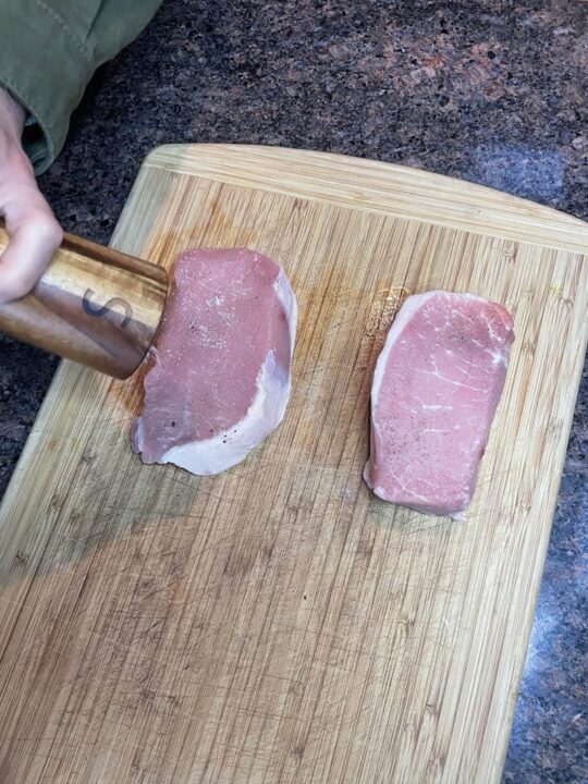 Two boneless pork chops on a cutting board being seasoned with salt.