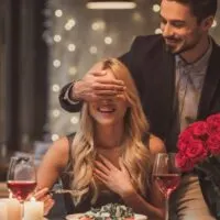 Man stands behind woman who's seated at table set for dining for two.